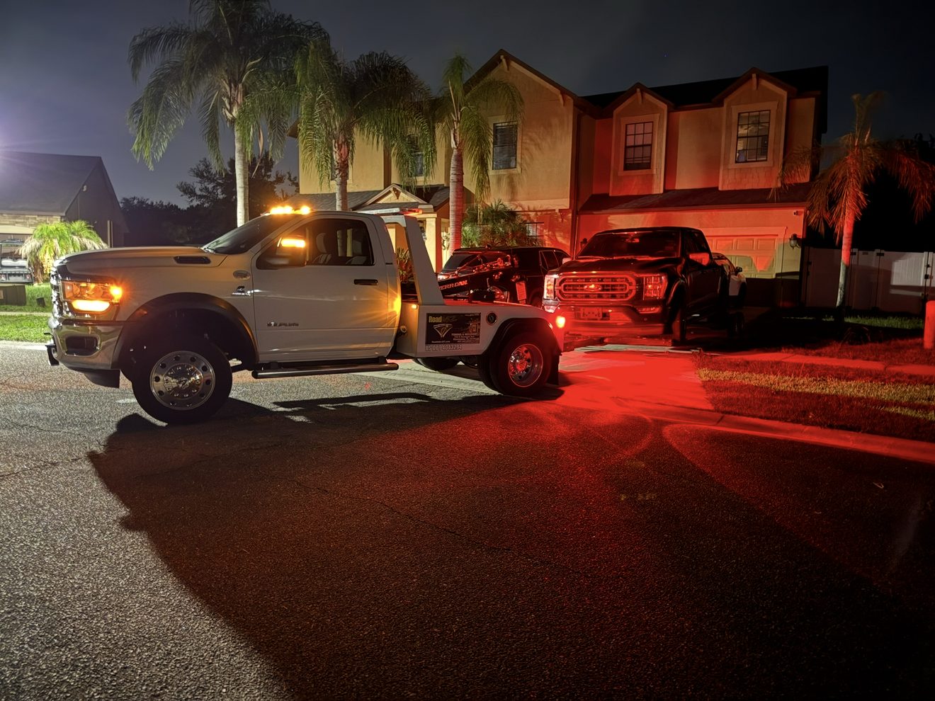 A tow truck performing a night-time recovery in a residential driveway with amber warning lights
